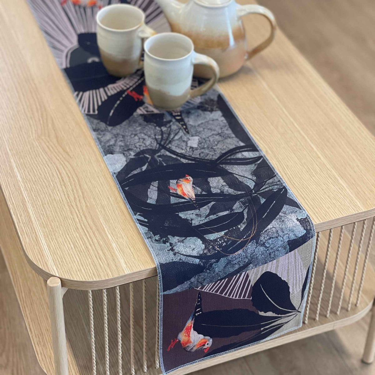 A rectangular table runner with a Japanese-inspired print of koi fish, leaves, and abstract patterns, styled on a light wood coffee table with ceramic mugs and a teapot.