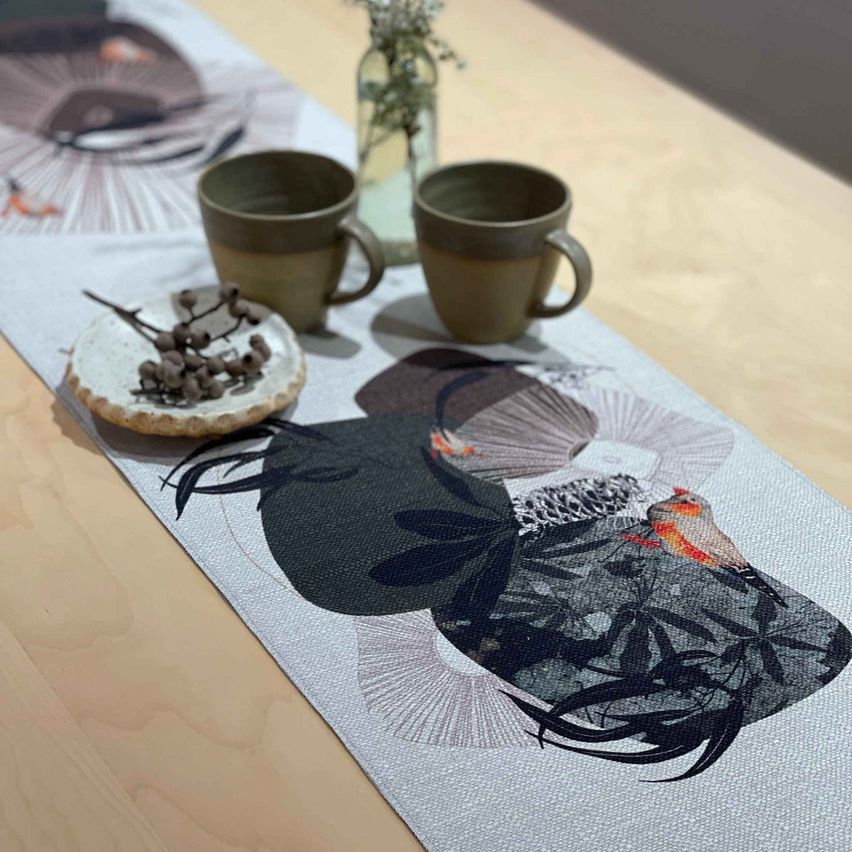 A light gray woven table runner with a black, gray, and orange bird and fan pattern, styled on a light wood table with two ceramic mugs, a small dish, and a glass vase.
